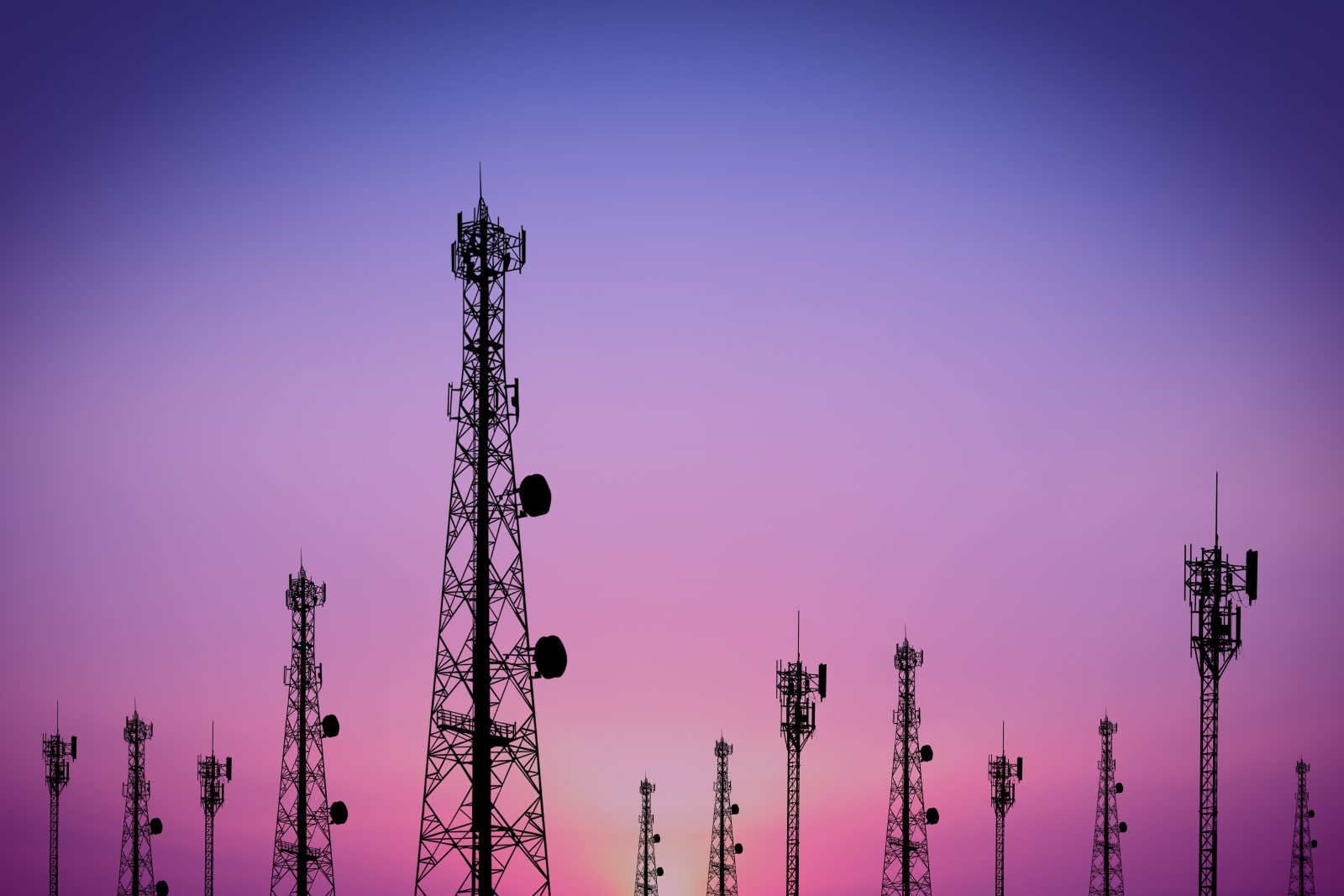 A field of cellular and telecom towers silhouetted against a purple twilight sky