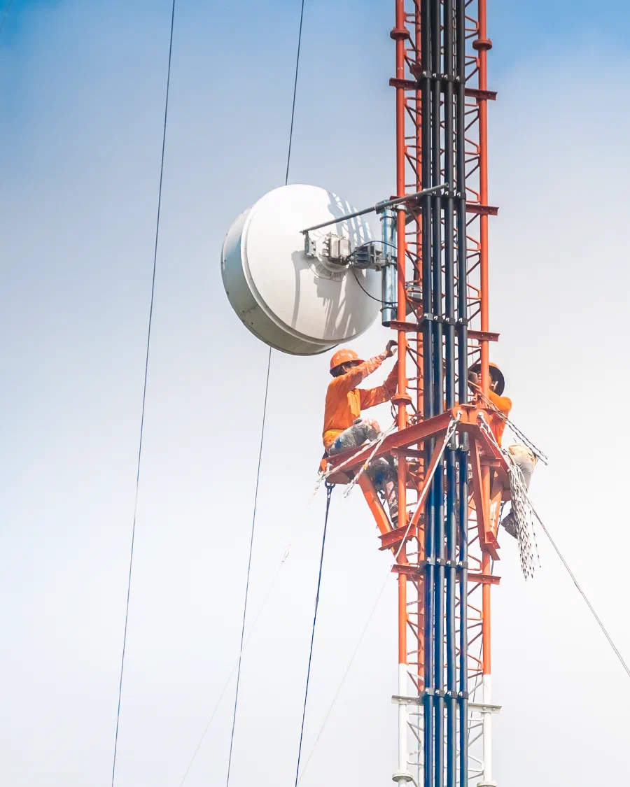 Two technicians working high on a telecom tower beside a microwave dish